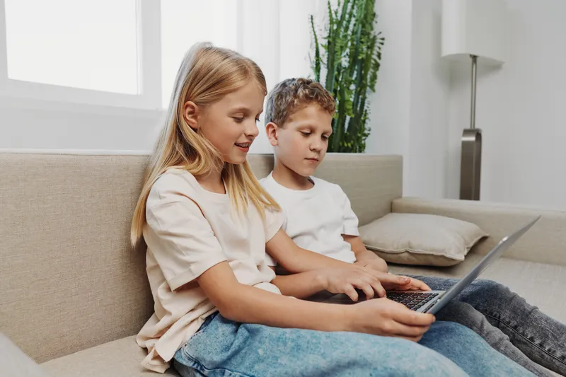 Two children sitting together on a sofa using a laptop, focused on screen in a bright modern living room with casual clothing and relaxed atmosphere.