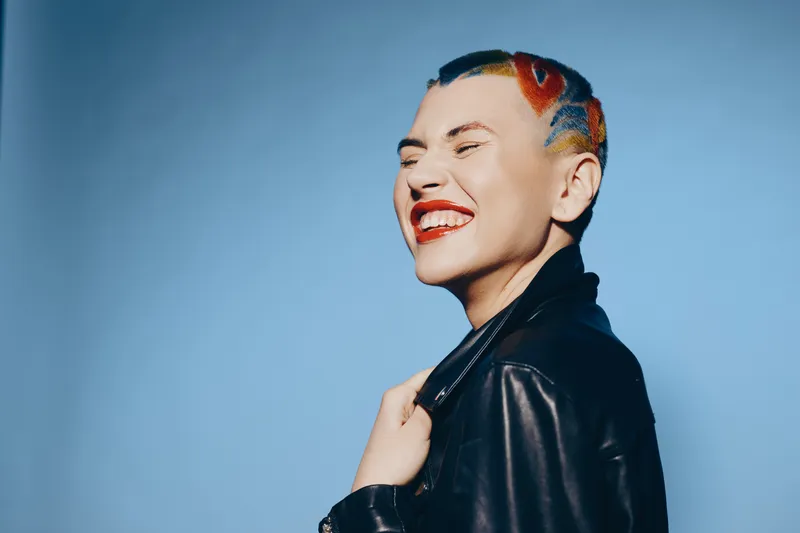 Young woman with colorful hair tattoos and bright makeup smiles joyfully wearing a black leather jacket against a clear blue background.