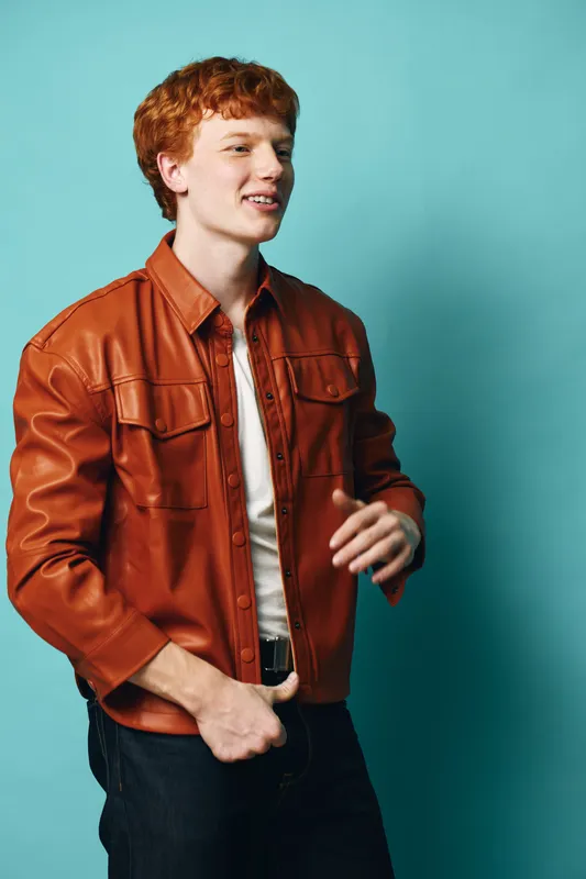 Young man with red hair wearing brown leather jacket and white shirt standing against blue background in casual style with confident expression and hand gesture.