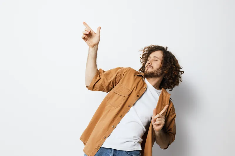Young man with curly hair in a casual outfit dancing joyfully against a plain background, showcasing emotions of happiness and freedom, ideal for lifestyle themes
