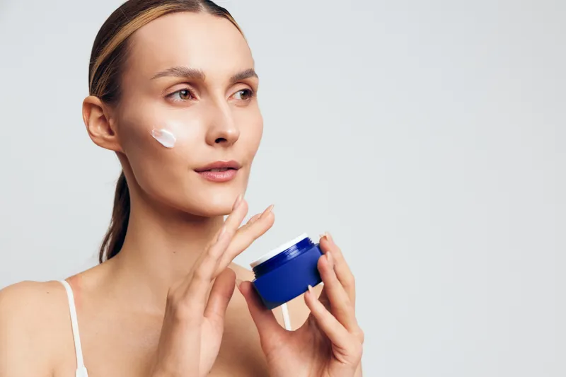Young woman with blonde hair applies moisturizing cream on her cheek while holding a blue jar against a plain light background, demonstrating skincare routine and self-care.