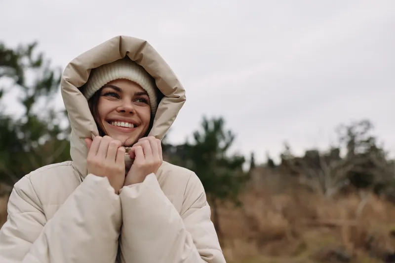 Woman in a warm beige jacket with a hood, smiling outdoors amid a chilly landscape, capturing a cozy moment in nature.