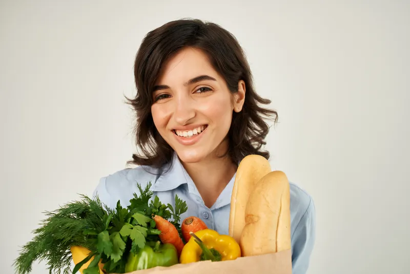 Улыбающаяся женщина в голубой рубашке держит сумку с продуктами и овощами, символизируя здоровое питание и органическую еду