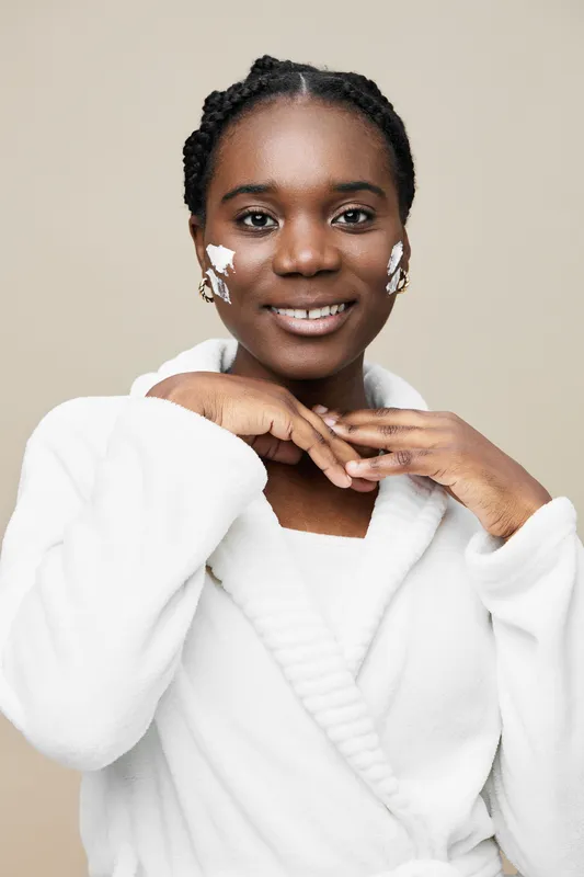 Smiling woman applying skincare on her face, wearing a cozy white robe against a beige background, embodying beauty, self care, and wellness, ideal for health related content