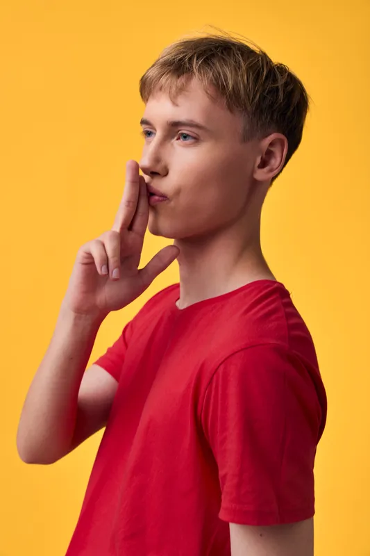 Shushing gesture by a young man in a red shirt against a bright yellow backdrop, conveying silence, secrecy and calm focus for creative stock use in a studio.