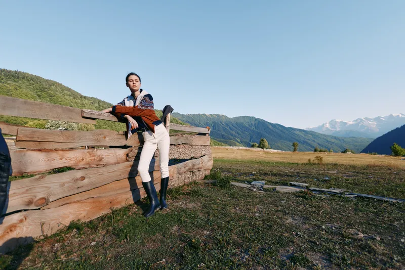 Woman leaning on wooden fence in countryside field with mountains in background, rural portrait outdoors wearing boots and casual jacket, scenic nature and open landscape.