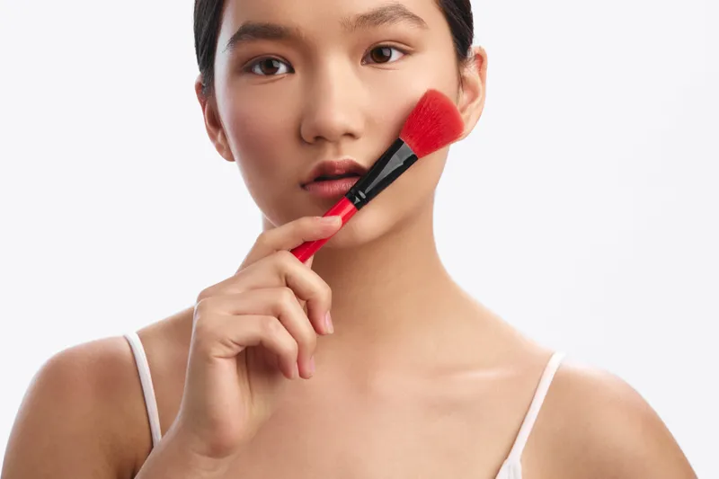 Asian woman applies makeup with a brush in a clean studio setting. Close up portrait capturing skin tone, focus on cosmetics, calm expression, subtle lighting, beauty routine, cosmetic tools, poised