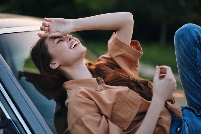 Joyful young woman laughing while leaning on a car hood, wearing a brown shirt and jeans, in a green outdoor setting, conveying happiness and relaxation.