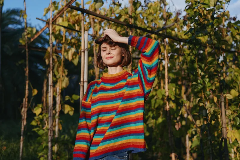 Young woman in her twenties wearing a striped sweater, smiling with eyes closed in tropical outdoor garden trellis, casual outfit and denim jeans, sunlight and relaxed vacation mood.