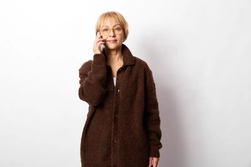elderly woman portrait in studio, thoughtful expression, finger raised near temple gesture, wearing brown fuzzy coat, blonde short hair, light background, calm mood