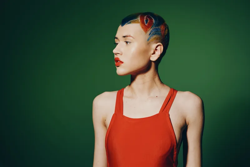 Woman with creative short hairstyle and red lipstick wearing a red sleeveless top against green background. Unique fashion portrait with bold color contrast.