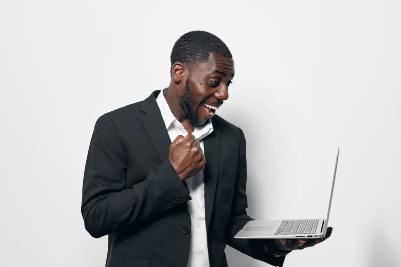 African American man with dark skin wearing black suit holding laptop and celebrating success with excited expression on white background in studio.