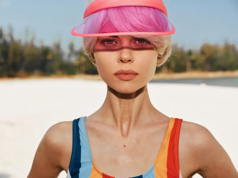 Close up portrait of a fashion forward model wearing a multicolor striped swimsuit and a pink visor, standing on a bright sandy beach under a clear blue sky.
