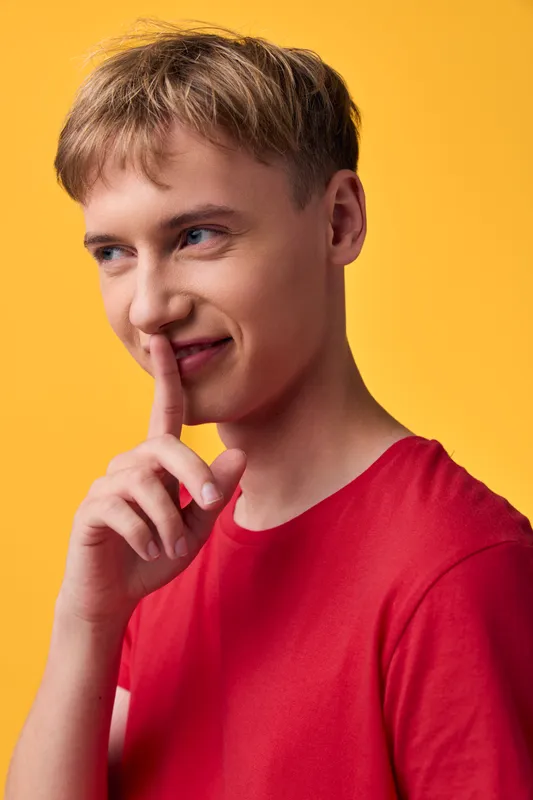 Thinking man with finger to lips in a casual pose against a bright yellow background, wearing a red shirt, conveying contemplation, curiosity and a lighthearted mood.