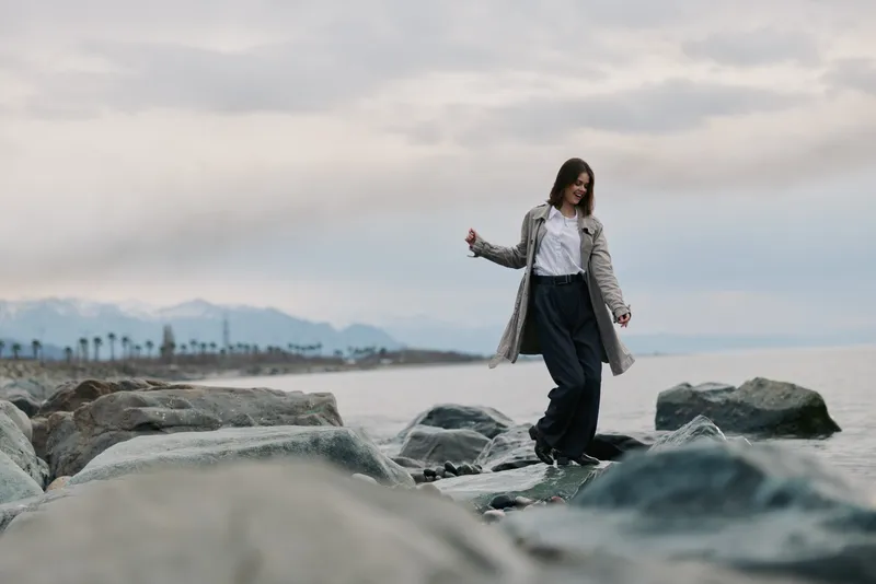 Young woman in casual outfit walking on rocks by the sea during cloudy day. Concept of nature, adventure, and calm lifestyle with mountains and water background.