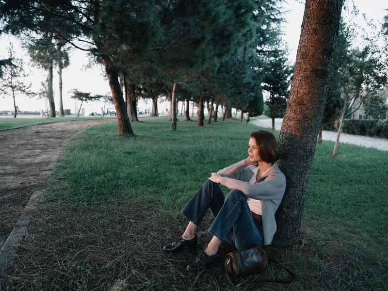 A contemplative woman sits beside a tree in a tranquil park. Leaning against the trunk, she wears casual clothes and gazes into the distance, creating a calm outdoor atmosphere and a moment of quiet