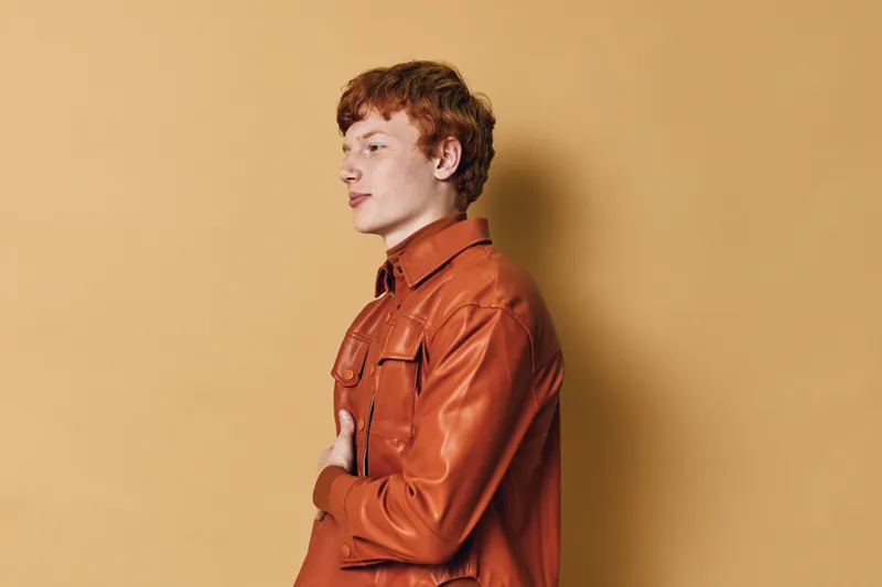 Young man with red hair wearing orange leather jacket posing sideways against beige background. Male model in casual stylish outerwear with thoughtful expression and relaxed posture.