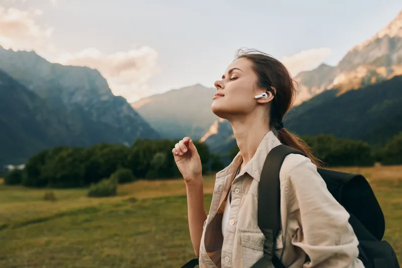 A young woman with a backpack stands in a mountain valley with her eyes closed, breathing in fresh air and savoring a peaceful moment amid soaring peaks and open space.