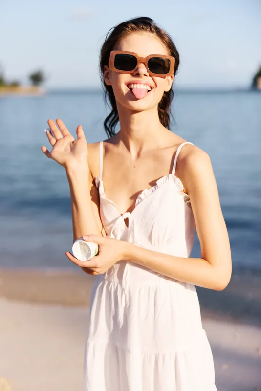 Young woman on a sunny beach posing with a shell, wearing a white sundress and oversized sunglasses, smiling and waving as calm sea and shore glow behind.