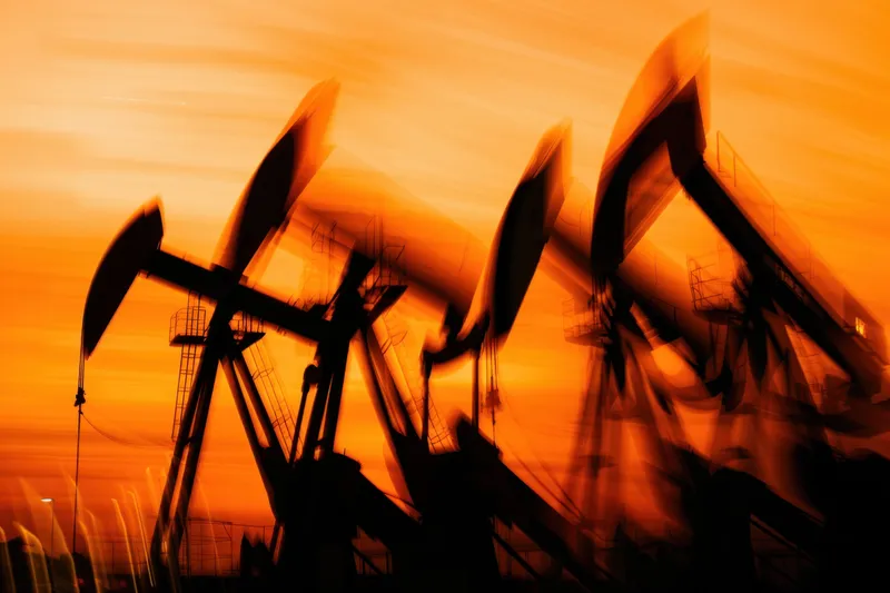 Абстрактное изображение нефтяных насосов в движении, силуэты которых выделяются на фоне драматического оранжевого заката