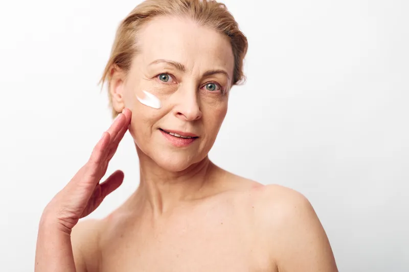 Mature woman applying cream on face with gentle smile on white background. Concept of skincare, aging skin care, self-care, beauty routine, and natural wellness.