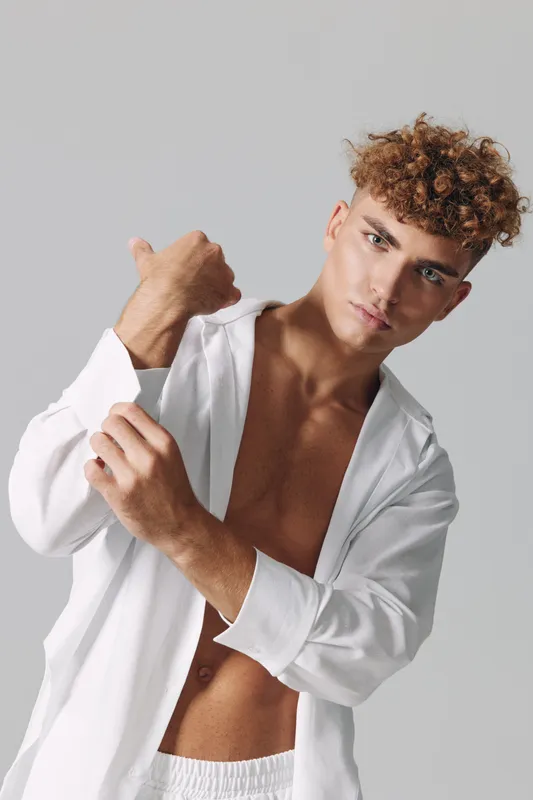Young man with curly hair wearing white open shirt and shorts posing against gray background in studio for fashion and lifestyle portrait photography.