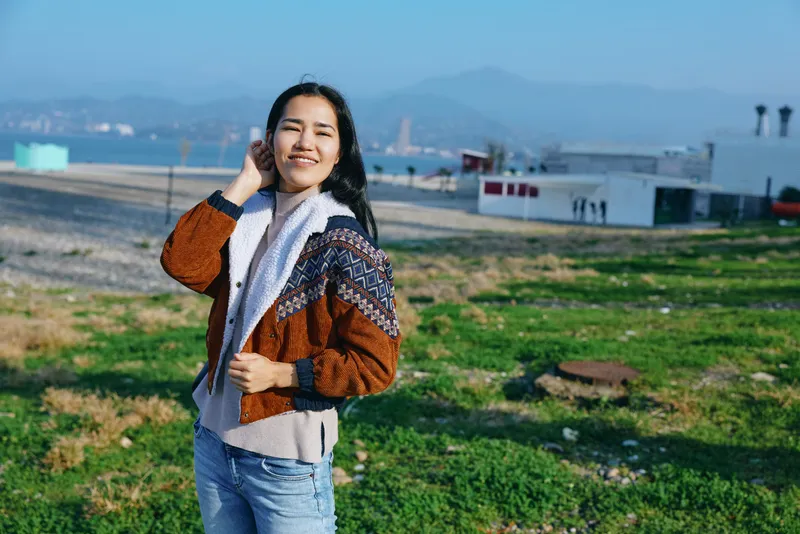 Woman in a patterned jacket and scarf standing in an open field, enjoying fresh air, casual pose, cheerful mood, bright day, distant buildings and blue sky, lifestyle photography for outdoor leisure