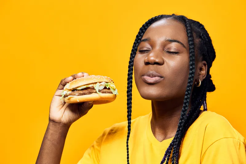 Young Black woman enjoying a delicious burger against a vibrant yellow background, showcasing joy and satisfaction in a casual style