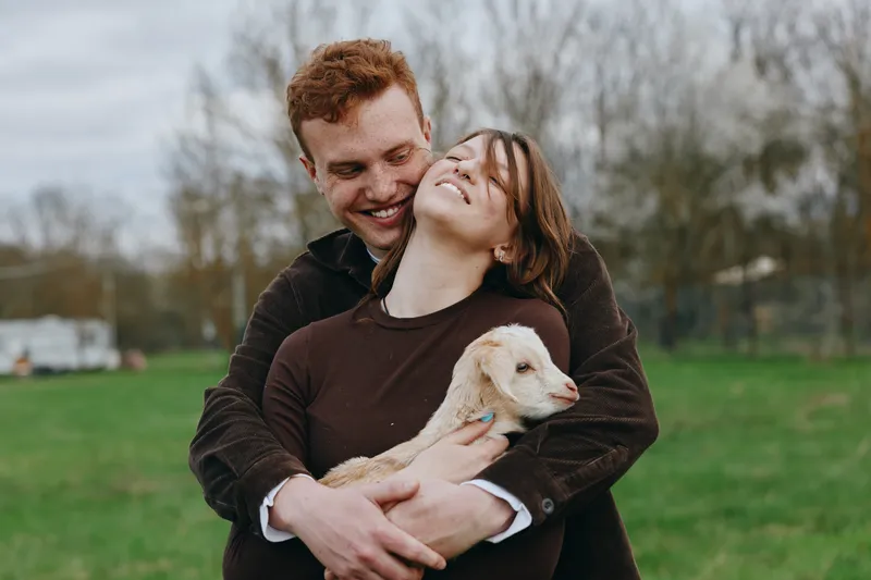 Joyful couple outdoors, embracing with a goat kid in a sunlit meadow, soft breeze, warm tones, candid moment, rustic setting, relaxed happiness, natural colors, peaceful countryside