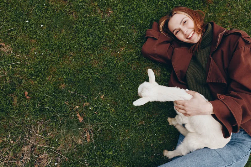 Woman with goat kid relaxing on grass outdoors, showcasing lifestyle and nature connection. Smiling, cozy pose, warm tones, soft daylight, serene countryside moment, gentle affection, simple