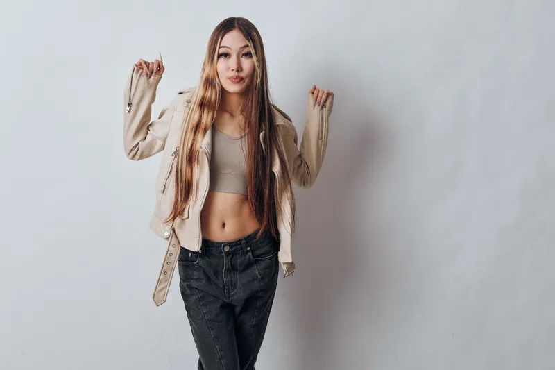 Young woman with long straight hair wearing casual beige jacket and crop top posing confidently against white background in studio. Fashionable girl with stylish outfit and relaxed expression.