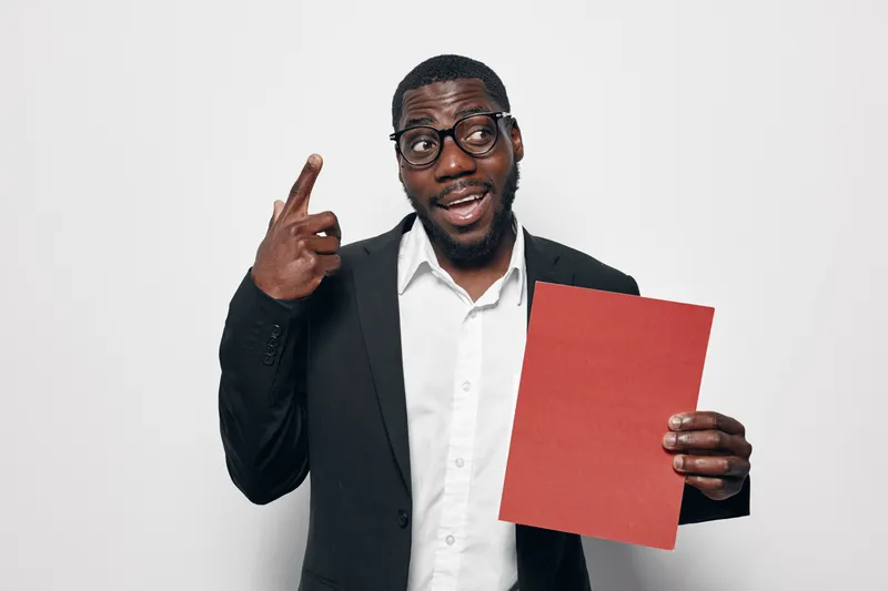 African American man with dark skin wearing glasses and black suit holding red folder and pointing finger up with thoughtful expression on white background.