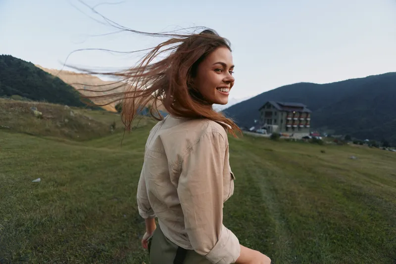 Smiling woman outdoors in a field turns toward the camera with wind in her hair. Casual clothing, natural light, and open landscape convey a carefree lifestyle and joy.