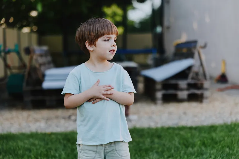 Young boy standing outdoors, displaying a thoughtful expression under the sunlight, wearing a light blue shirt and light pants, amidst a playful background.