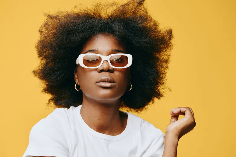 Stylish young woman with natural afro hairstyle, wearing white sunglasses and a minimalist t shirt against a vibrant yellow background, showcasing confidence and individuality