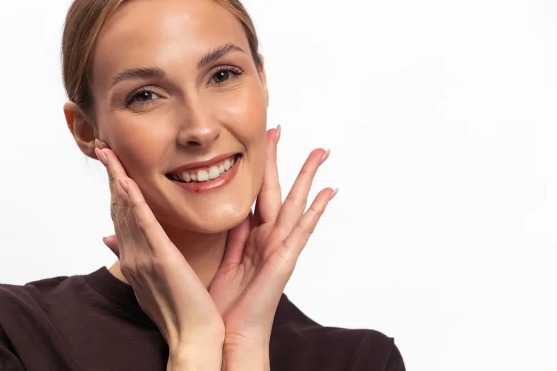 Smiling young woman with clear skin touches her face gently, wearing a dark top on white background. Concept for beauty, skincare, health, and natural appearance.