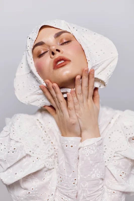 Elegant young woman in a white vintage dress and bonnet, showcasing soft emotions against a minimalistic gray background