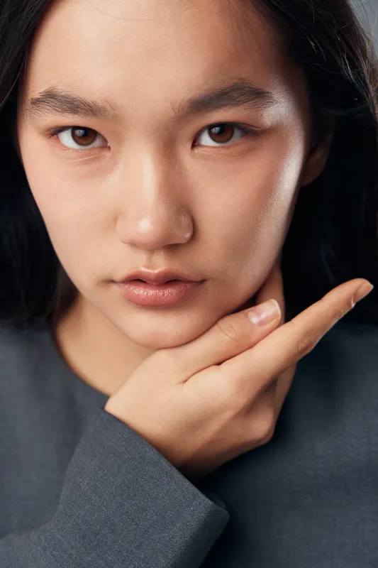 Portrait of young woman with natural skin and serious expression touching chin in closeup. Female model with dark hair and soft lighting looking directly into camera lens.