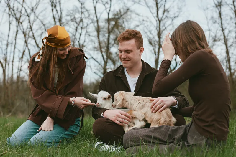 A candid outdoor scene shows a goat kid being petted by three adults sitting on grass, conveying family bonding, gentle care, and the simple joys of rural life.