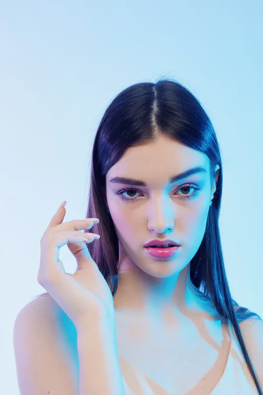 Young woman beauty fashion model with long dark hair posing with hand near face. Studio portrait with blue lighting on white background. Concept of elegance and style.