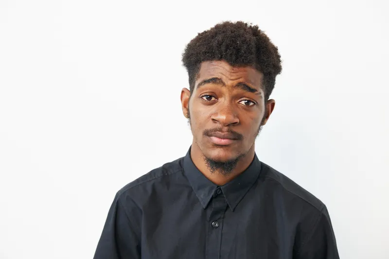 A young African American man expressing contemplation, dressed in a black shirt, showcasing a thoughtful demeanor against a minimalist background.