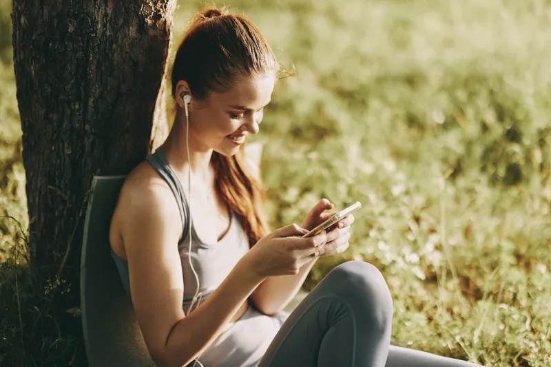 Young woman enjoying music on her smartphone while sitting outdoors, wearing casual activewear, showcasing relaxation and happiness in a sunny park setting