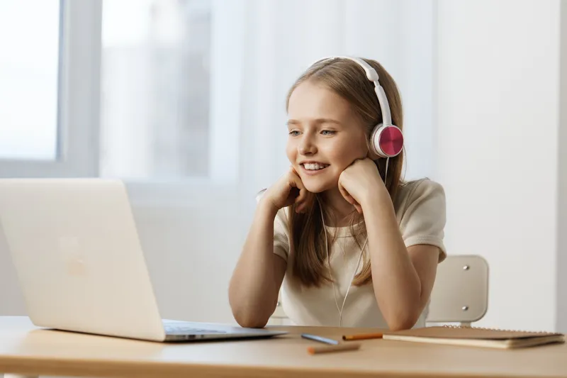 Smiling girl with headphones enjoying online learning at home, bright atmosphere, cozy interior with a laptop and stationery elements