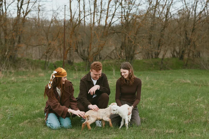 Three young adults enjoy a spring afternoon outdoors while playing with two baby goats on a green grassy field near leafless trees.