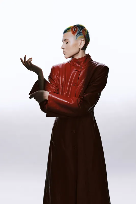 Artistic woman with colorful hair wearing a long red leather coat, posing elegantly against a minimalistic white background with soft lighting highlights.