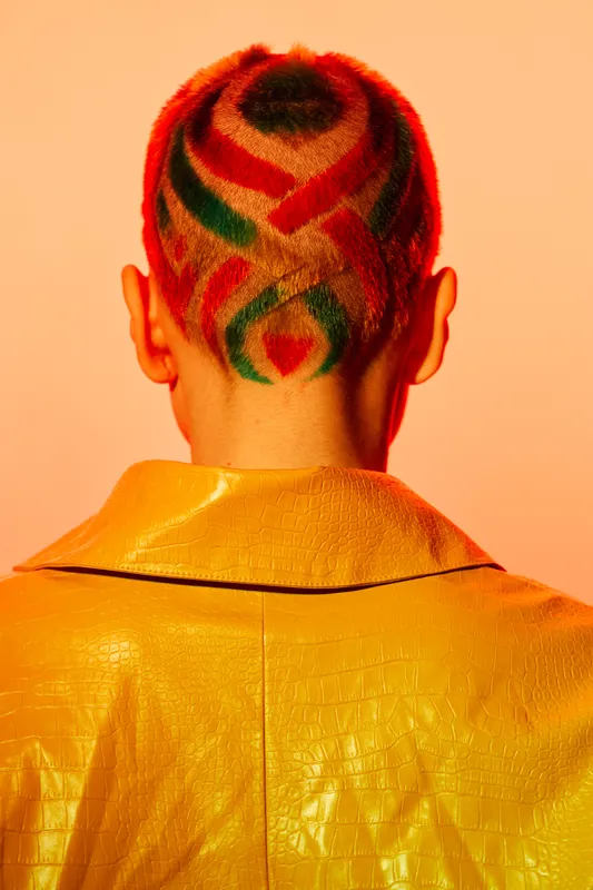 Woman with colorful geometric shaved haircut wearing a textured yellow jacket, standing against a warm orange background, showcasing creative hairstyle and bold fashion.