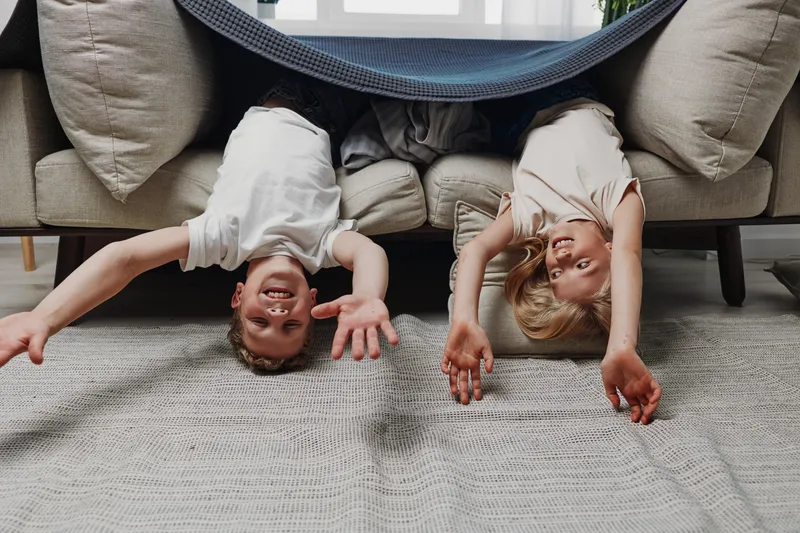 Two happy children hang upside down from a couch, smiling and reaching toward the camera in a cozy living room. Playful mood and family fun captured indoors with natural light.