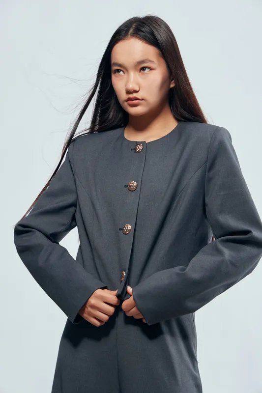 Young woman with long dark hair wearing a gray tailored jacket with buttons standing against light background looking away thoughtfully with hands adjusting the coat for fashion portrait.