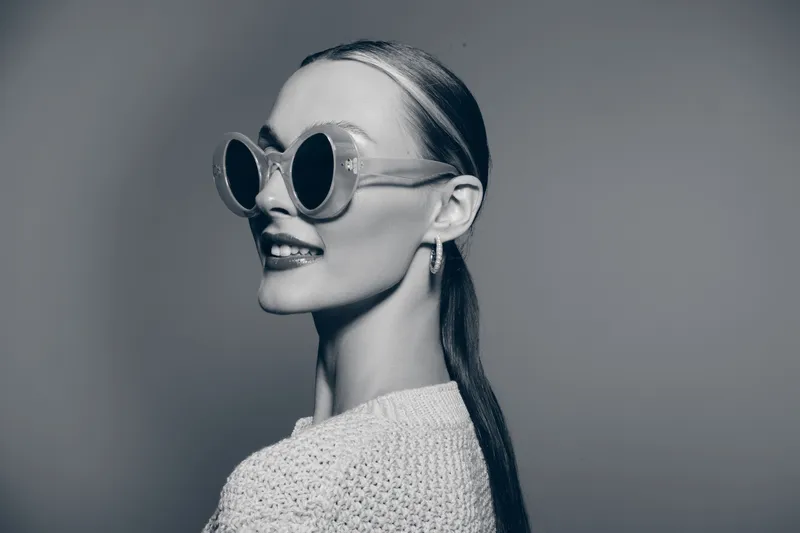 Stylish Caucasian woman with fair skin and pulled-back hair poses wearing round sunglasses and a knitted sweater in a monochrome studio setting.