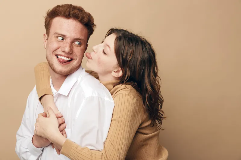Happy young couple embraces and smiles in casual clothes on beige background. Woman hugs man from behind and looks at him with affection. Concept of love, relationship, and intimacy.